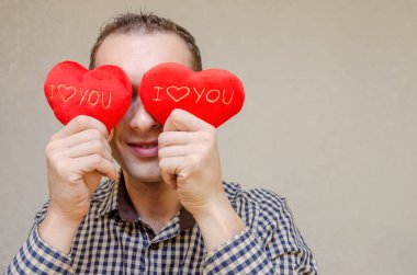 Elinde iki küçük kırmızı kalple gülümseyen koyu renk saçlı çekici bir adam gözlerini kağıt kalp sembolleriyle kaplıyor, açık bir arkaplanda duruyor. Sevgililer Günü