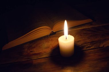 Tahta bir masanın üzerinde, karanlıkta yanan bir mum ve bir kitap. Bilgi, din, sihir kavramı