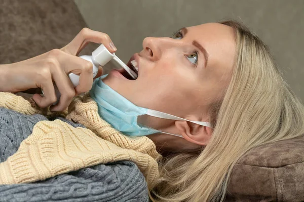 young blonde woman with a scarf around her neck and a medical mask pulled down on her chin is lying on the sofa and stabbing her throat with a spray