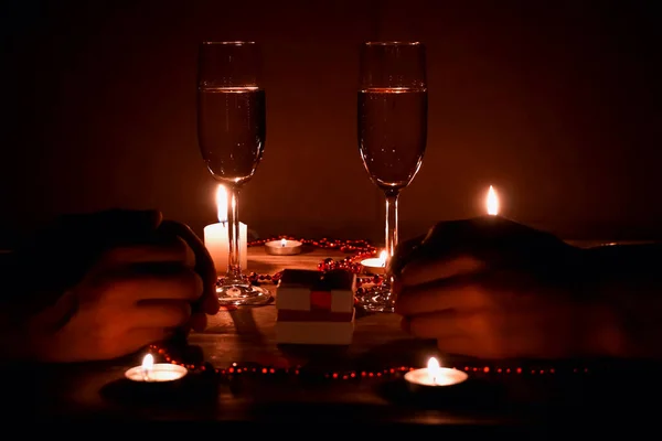 Close-up of a pair of hands on a table with two glasses of champagne, candles, hearts, beads. A romantic couple hold each other\'s hand during dinner. concept of engagement.