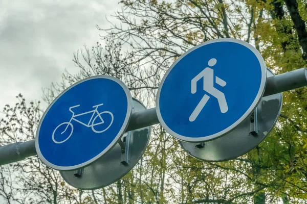 Two road signs side by side for pedestrians and cyclists close up against the sky and tree branches