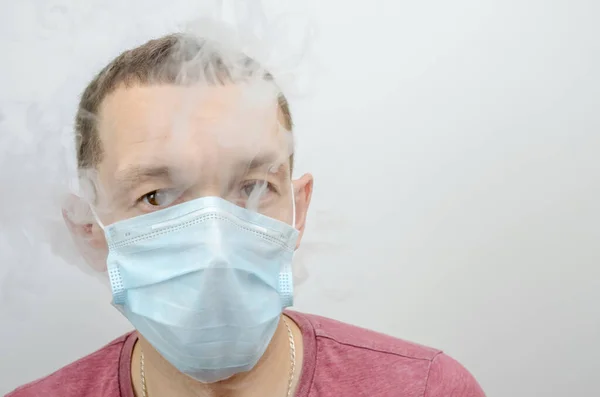 man lets out smoke from under a medical mask on his face on a light grey background