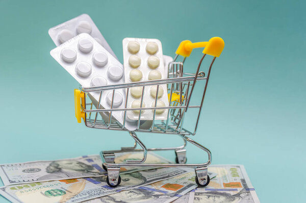 Желтая тележка с таблетками стоит на долларовых купюрах на синем фоне крупным планом. Концепция здравоохранения, интернет-магазины, высокая стоимость лекарств