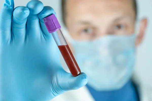 Male doctor in a medical gown, mask and gloves holds a test tube with blood close up. Concept of medical tests