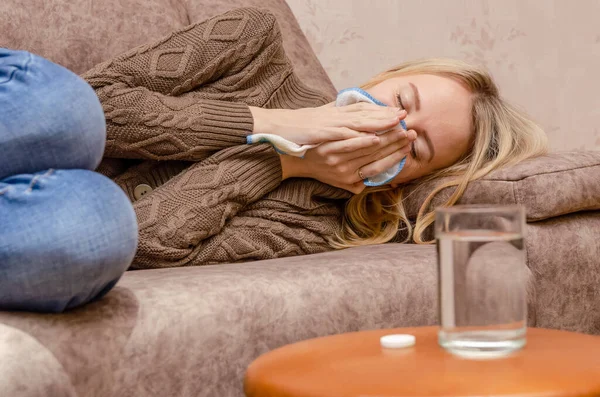 blonde girl sitting on the couch. She has a runny nose, so she blows her nose and wipes away tissue paper. place the medicine and a glass of water on the table next to it. The concept of the disease