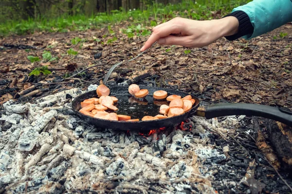Sausages are fried in a pan on coals in the forest. The girl\'s hand turns the sausage with a fork in a pan on the coals. Breakfast in nature. Picnic.