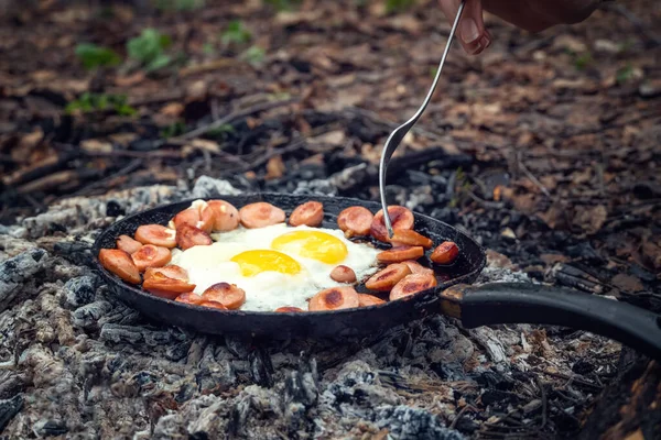 Girl\'s hand with a fork turns over pieces of fried sausage and eggs in a pan on the coals. Breakfast in nature, picnic, camping food.