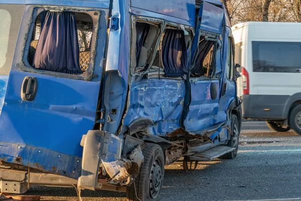 A rumpled blue minibus is on the road after an accident. Accident on the road in the city on a sunny day. A truck crashed into a minibus and crashed a car. Broken blue minivan close up