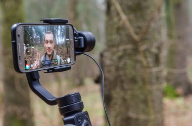 Erkek blogcu kendini ormanda cep telefonu kamerasıyla çekiyor ve kameraya gülümsüyor ve el sallıyor. Blogculuk, internet, bilgi