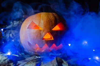 Parlayan gözlü turuncu balkabağı, dumandaki mavi ışıkta karanlıkta bir ağız. Cadılar Bayramı konsepti.