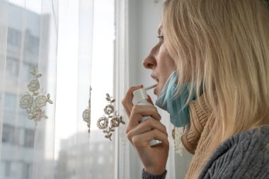Çenesinde maske olan sarışın kız pencerenin yanında dururken boğaz spreyi sıkıyor. Sağlık ve Tıp Konsepti.