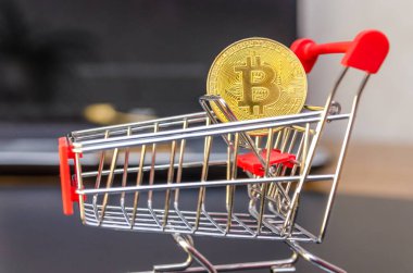 Kırmızı bir alışveriş arabasındaki altın bitcoin siyah bir arkaplanda dizüstü bilgisayarın önünde duruyor.