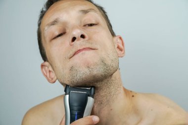 Açık arkaplanda elektrikli jileti olan koyu renk saçlı güzel bir adamın portresi. Koyu renk saçlı adam yakın plan elektrikli tıraş makinesiyle tıraş oluyor..