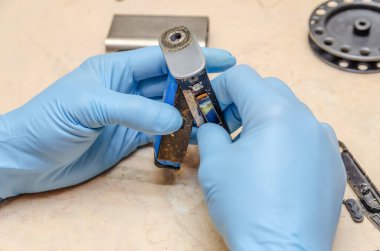 Mavi eldivenli bir erkek, ahşap bir masada elektronik bir sigarayı tamir ediyor.