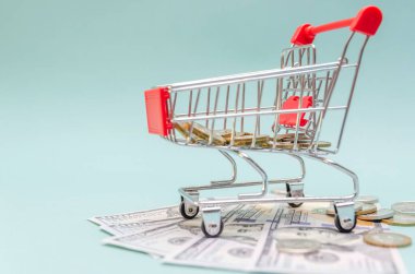 Altın ve gümüş sikkeli kırmızı alışveriş arabası mavi arka planda dolar banknotlarının üzerinde duruyor. Çevrimiçi alışveriş, online alışveriş