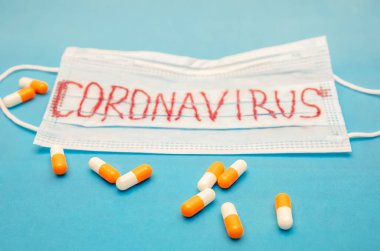 Üzerinde koronavirüs yazan tıbbi maske ve mavi arka planda dağınık turuncu ve beyaz kapsüller.