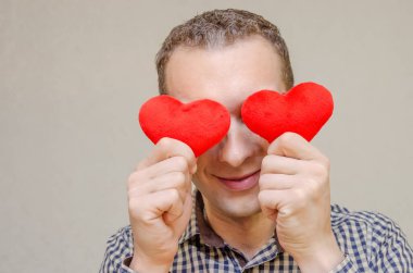 Elinde iki küçük kırmızı kalple gülümseyen koyu renk saçlı çekici bir adam gözlerini kağıt kalp sembolleriyle kaplıyor, açık bir arkaplanda duruyor. Sevgililer Günü