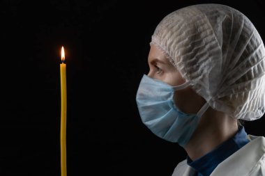 Genç bir kadın tıbbi bir maske, tıbbi bir başlık ve beyaz önlük takmış elinde yanan bir mumla siyah bir arka planda dikiliyor. Parlak bir geleceğe olan inanç kavramı.