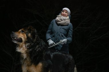 Bir kış gecesinde parkta yürüyen Alman çoban köpeği olan bir kız. Karanlık bir ormanın arka planında fotoğraf.