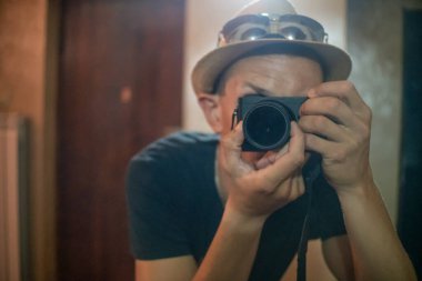 Hasır şapkalı ve gözlüklü genç bir adam fotoğraf makinesi merceğine odaklanmış aynanın içinden selfie çekiyor..