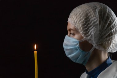 Genç bir kadın tıbbi bir maske, tıbbi bir başlık ve beyaz önlük takmış elinde yanan bir mumla siyah bir arka planda dikiliyor. Parlak bir geleceğe olan inanç kavramı.