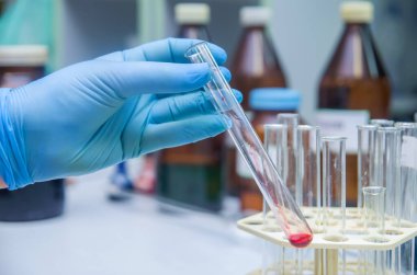 Bir adamın el ele tutuşması ve bir cam test tüpünü (Proberohr) labaratuvardaki yansıyan masanın üzerine kırmızı (sıvı, morumsu) suyla yaslaması.