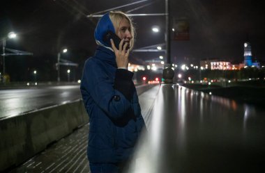 Mavi ceketli ve kafasında atkısı olan genç bir kadın köprüde arabalarla cep telefonuyla konuşuyor.