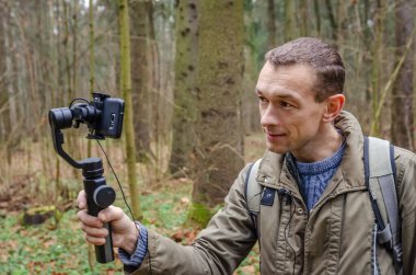 Erkek blogcu kendini ormanda cep telefonu kamerasıyla çekiyor ve kameraya gülümsüyor ve el sallıyor. Blogculuk, internet, bilgi