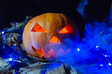 Parlayan gözlü turuncu balkabağı, dumandaki mavi ışıkta karanlıkta bir ağız. Cadılar Bayramı konsepti.