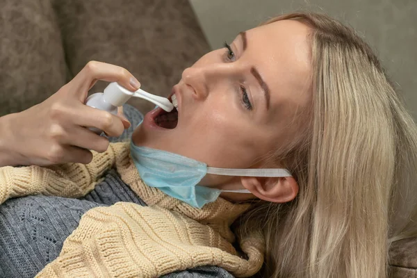 young blonde woman with a scarf around her neck and a medical mask pulled down on her chin is lying on the sofa and stabbing her throat with a spray