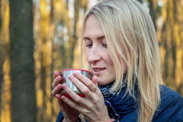 Genç sarışın bir kız gün batımında bahar ormanında termos bardağından çay içer. Tema yolculuğu. Yürüyüş sırasında çay içen kız.