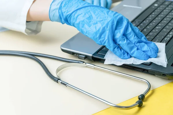 woman\'s hand in blue medical gloves wipes the laptop keyboard with a disinfecting cloth, and a stethoscope is lying next to it