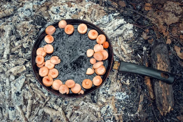 Sausages are fried in a pan on coals in the forest. Breakfast in nature. Picnic.