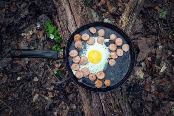 Frying pan with eggs, sausage, and a piece of bread stands on a fallen tree in the forest, top view. Concept of tourism and travel. Breakfast in nature