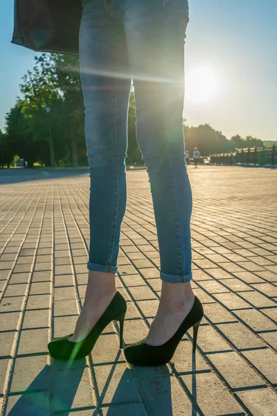 Slim female legs with blue jeans and black suede high-heeled shoes against the background of the sun at sunset. Stylish elegant girl walking in the park. vertical photo