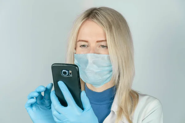young female doctor in a mask and white coat uses a smartphone on a light background with copy space