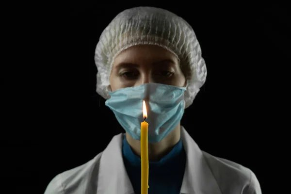 Young woman in a medical mask, medical cap and white coat holds a burning candle in front of her on a black background. Concept of last hope, faith in a bright future.