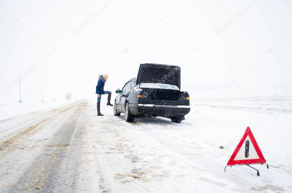 woman changing the wheel of a black car with an open trunk in winter snow with the alert sign on a snowy road