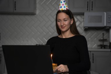 Genç bir kadın doğum gününü internette kutluyor, şenlikli bir parti şapkası takıyor ve mutfağında üzerinde yanan bir mumla oturmuş, neşeli bir anın tadını çıkarıyor.
