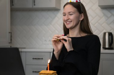 Parti şapkası takan gülümseyen genç bir kadın, doğum gününü mutfağında, önünde bir doğum günü pastası ve yanan bir mumla kutlarken parti düdüğü çalıyor.