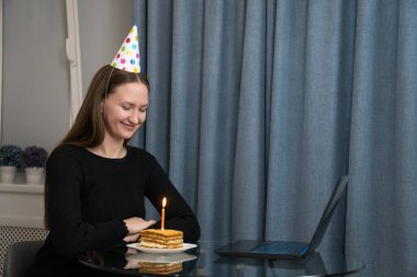 Parti şapkası takan genç bir kadın bilgisayarının önünde doğum günü pastası ve mumla doğum gününü internette kutluyor.