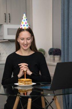 Doğum günü pastası ve mumla cam masada oturan parti şapkası takan genç bir kadın online doğum günü partisi kutlaması sırasında dilek tutuyor. Dikey fotoğraf