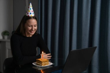 İnternette doğum gününü kutlayan parti şapkası takan genç bir kadın dizüstü bilgisayarla, mumla pasta parçasına bakıyor arkadaşları ve ailesiyle video görüşmesi yapıyor.