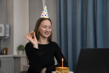 Parti şapkası takan genç bir kadın internetten doğum gününü kutluyor. Parti kornası çalıyor ve evdeki video görüşmesi sırasında mum ışığında pastaya bakıyor.