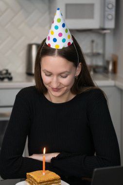 Genç bir kadın doğum gününü internette kutluyor, şenlikli bir parti şapkası takıyor ve mumla lezzetli bir pastanın tadını çıkarıyor, rahat mutfağında dilek tutuyor. Dikey fotoğraf
