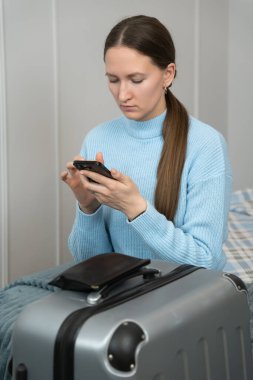 Genç bir kadın, bir otel odasında bavulunu toplarken akıllı telefonunu kullanıyor. Gidişine ya da gelişine hazırlanıyor. Seyahat düzenlemelerini yapıyor ya da diğerleriyle iletişim kuruyor. Dikey fotoğraf