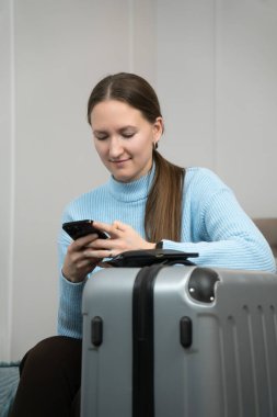 Genç bir kadın, akıllı telefonuyla eve uçmayı bekliyor. Pasaportu ve bavulu yakınlarda otururken seyahat ayarlamaları yapıyor. Yaklaşan yolculuk için heyecan saçıyor. Dikey fotoğraf