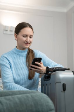 Akıllı telefon tutan genç bir kadın, rahat bir otel odasında ya da evde rahat bir kanepede dinlenirken yaklaşan tatilini düşlüyor. Dikey fotoğraf
