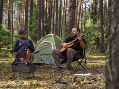Happy couple enjoying a serene camping experience in the forest, sitting by a warm bonfire, strumming a guitar, and unwinding in nature near their cozy tent