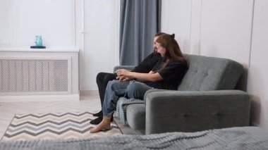 A girl and a man are sitting on a comfortable sofa in their living room, having a deep conversation that seems to cause the woman suffering and sadness